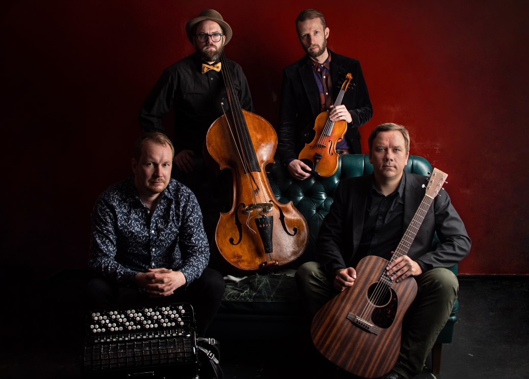 Gruppenaufnahme von 4 Musikern mit ihren Instrumenten vor rotem Vorhang
