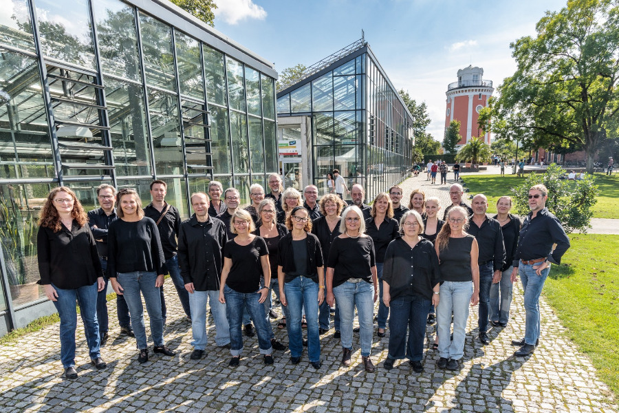 27-köpfiger Chor -schwarzes Oberteil, blaue Hose- vor einem Gewächshaus